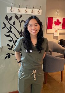 Smiling Asian woman in olive scrubs stands in a clinic lobby with a plant mural and a Canadian flag on the wall behind her.