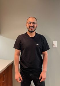 Smiling man in black scrubs standing in a clinical room with a light gray wall behind him.
